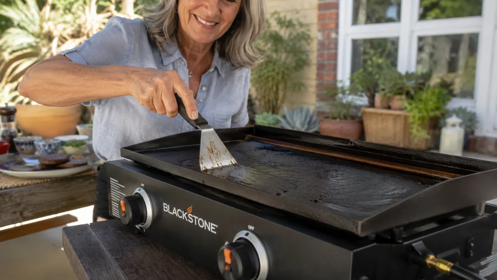 Close-up of hands scraping a Blackstone griddle surface clean with a metal scraper, grease trough catching runoff